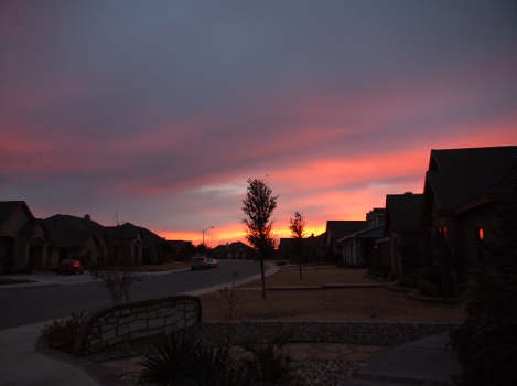 [A swath of yellow dashes across the sky close to the horizon with swathes of pink on either side of it. Rising into the sky are swatches of peachy-pink and purple. The peach color is reflected on a house window in the foreground.]